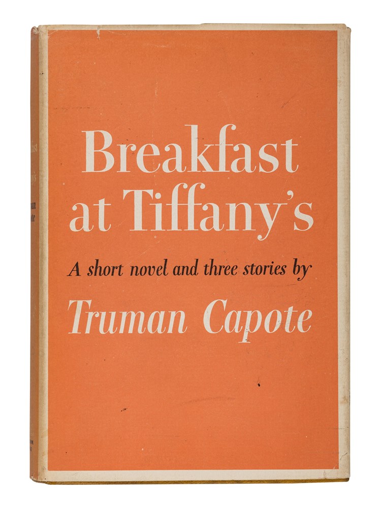 A photo of the novel Breakfast at Tiffany's by Truman Capote and it's clamshell covering. The novel is facing straight on, in it's original orange dust jacket. To it's left is the clamshell case, angled at 45 degrees. The clamshell is 1/4 black leather and 3/4 orange cloth, featuring an inset of Audrey Hepburn on the cover. 